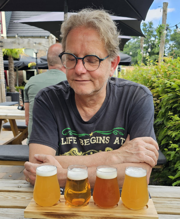 Man with blond hair and glasses sits at an outdoor table with a flight of four beers in small glasses in front of him.