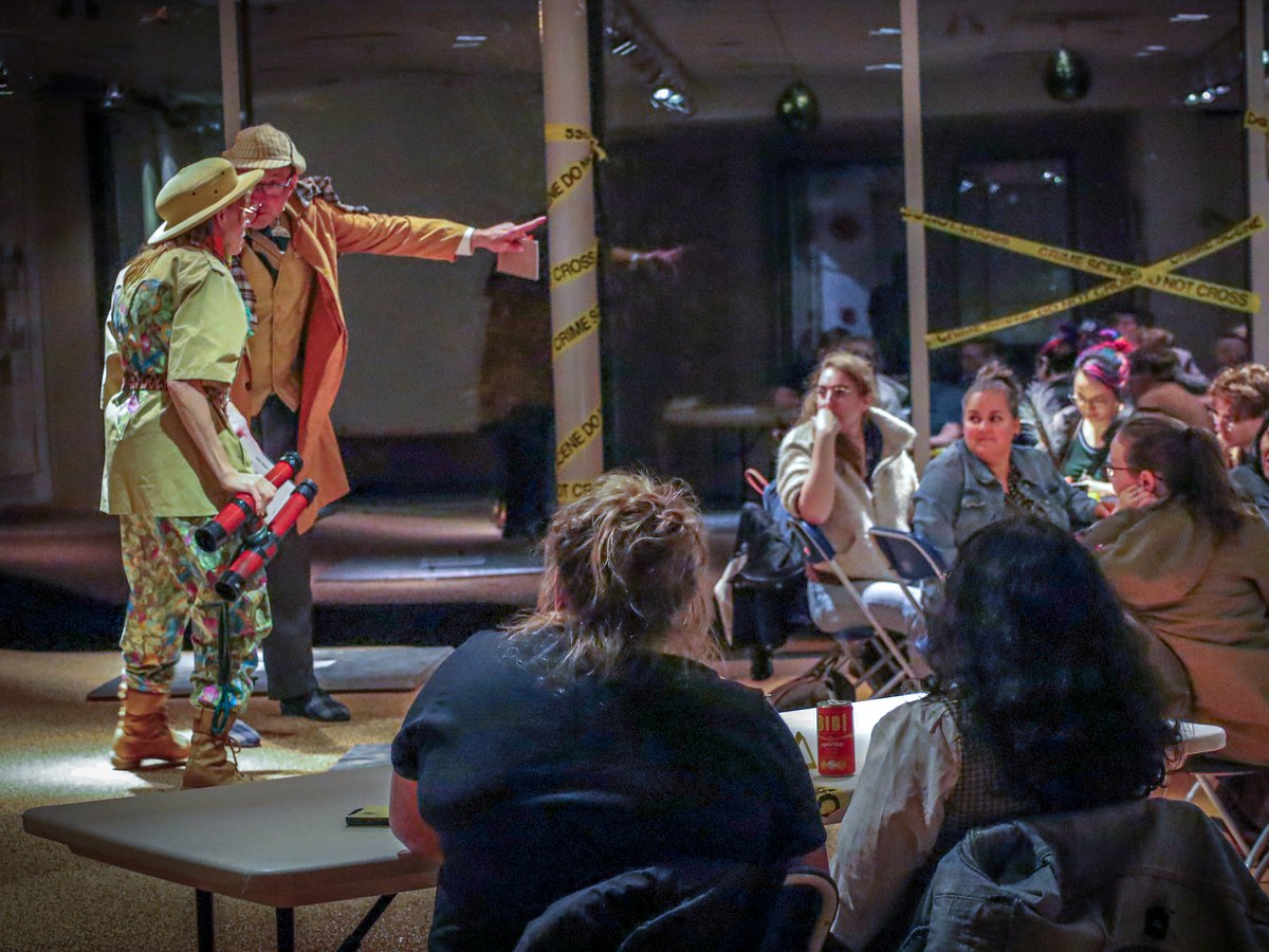 The detective naming the killer beside a costumed suspect as guests watch at a Murder YOU Mystery at the Museum of Vancouver