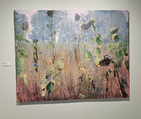 Nature's Secret Game II by Isabella Werkhoven. A large painting of a field with tall, wilting sunflowers and wildflowers in muted tones of green, brown, and blue. The background features a hazy sky and distant trees, creating a moody, atmospheric effect. The painting hangs on a light gray gallery wall with a small label beside it.