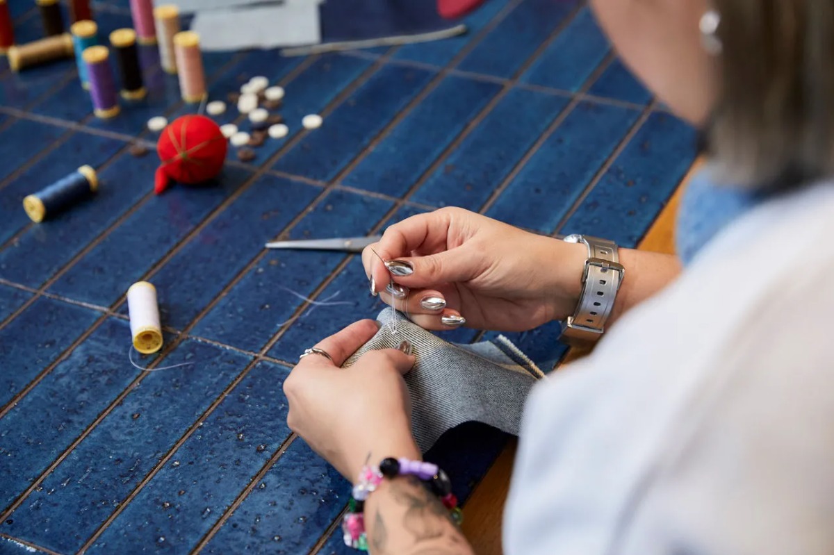 Close-up of a person's hands hand-sewing a piece of denim fabric at a blue tiled table.
