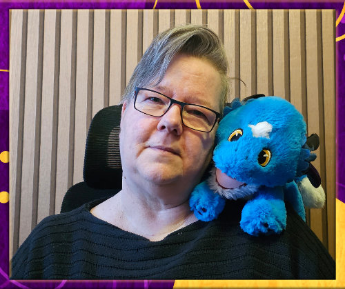 Blaine—short greying hair and glasses—sits in front of a wooden panel wallpaper, smiling gently. A bright blue plush dragon with a white spot on its forehead rests on eir shoulder. The image has a purple and gold border.